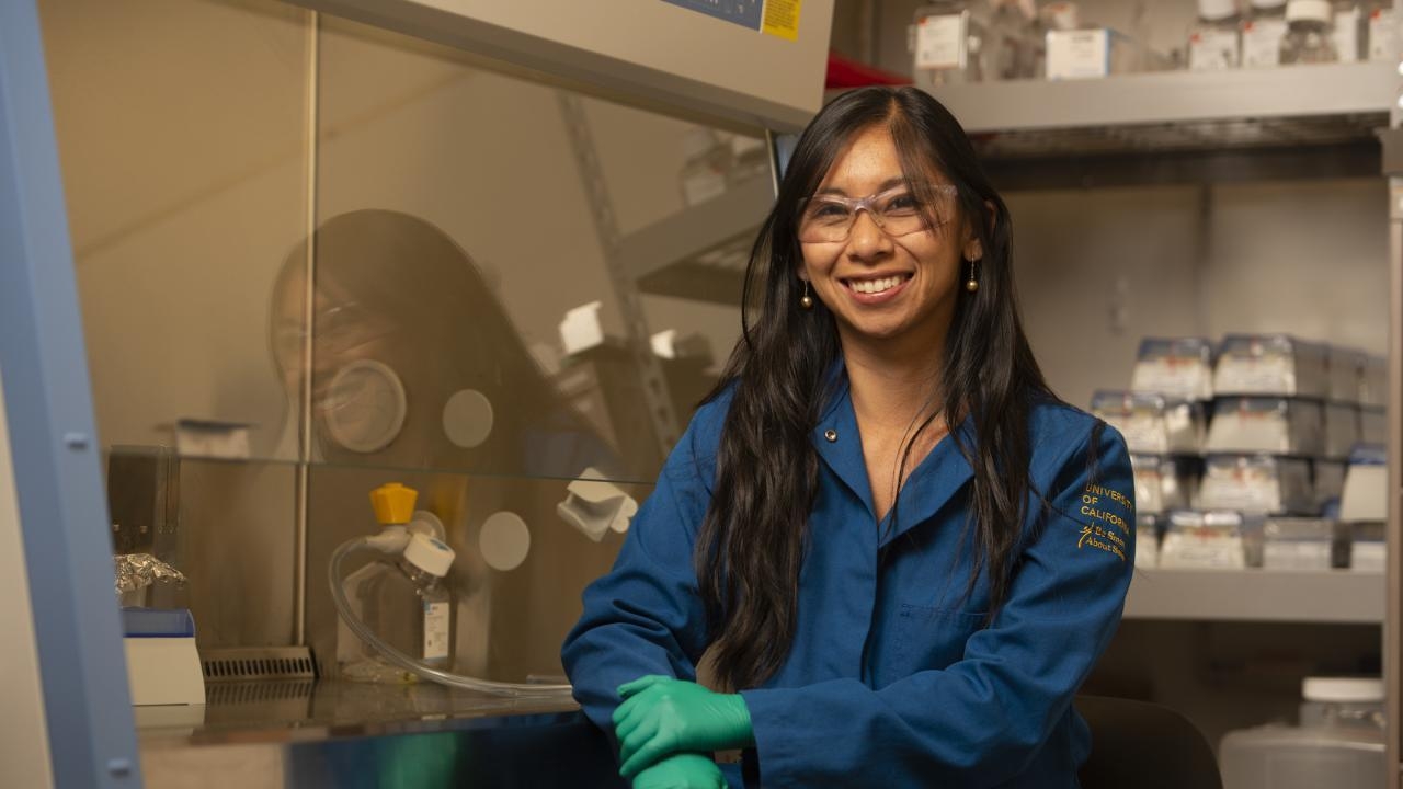 Marie Heffen in a blue lab coat sitting in a lab and smiling at camera